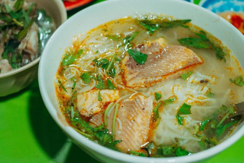 Semangkuk ikan kuah kuning berbumbu rempah dengan potongan ikan kakap, tomat hijau, belimbing wuluh, daun kemangi, dan taburan kenari siap disajikan hangat.